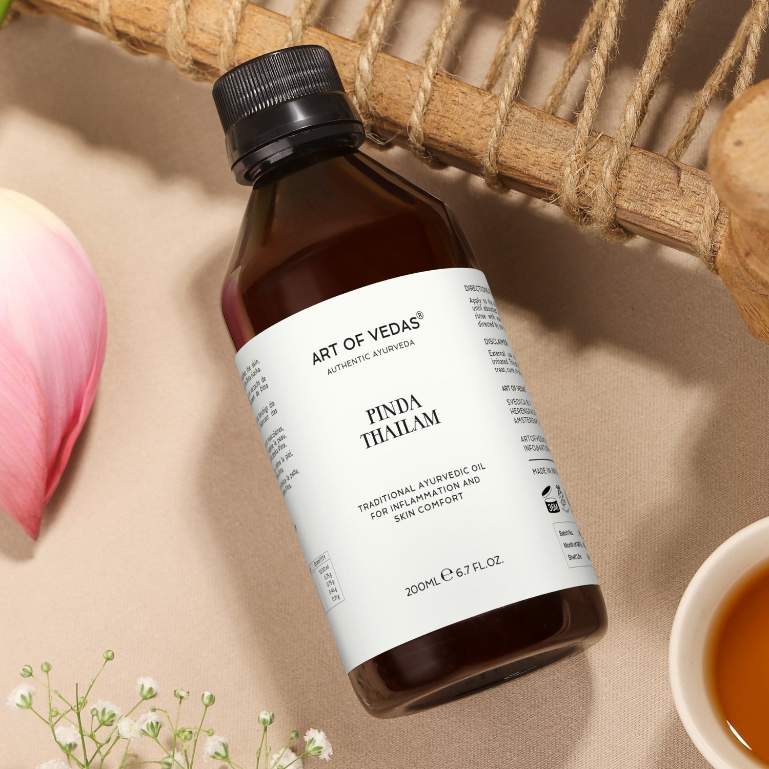 Bottle of Art of Veda Pinda Thailam oil on a beige surface with a pink flower and small bowl.