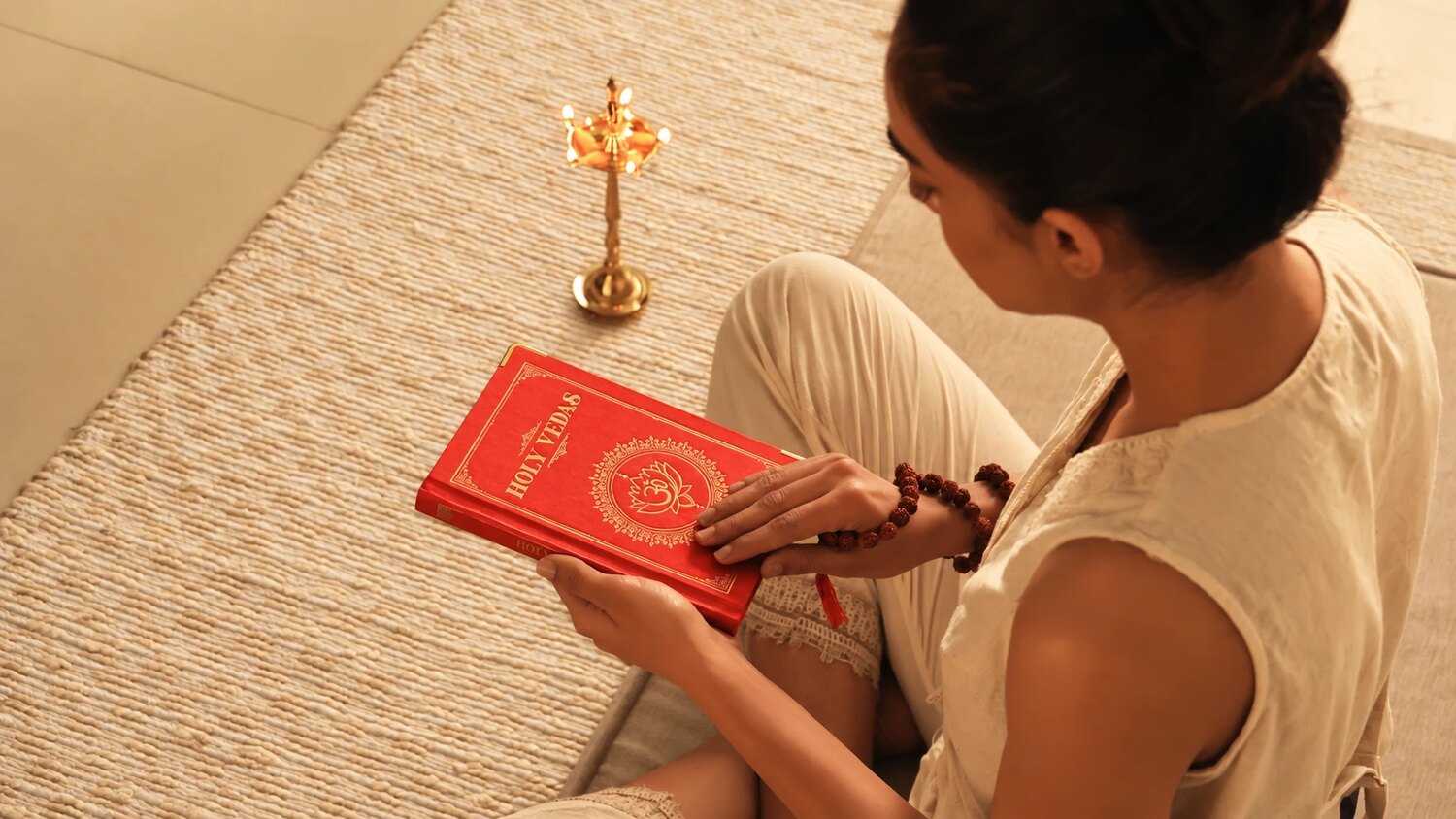 Mujer sentada sobre una esterilla leyendo un libro rojo de los Vedas sagrados junto a una lámpara ayurvédica de latón, para Art of Vedas